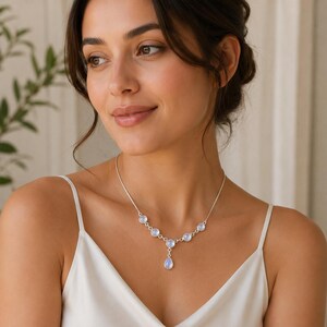 May include: A silver necklace with a series of iridescent moonstone gems and a teardrop pendant. The necklace is worn by a person wearing a white top. The jewellery is displayed against a soft, neutral background.