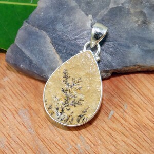 May include: A teardrop-shaped pendant with a silver bail and a beige stone with black dendritic inclusions.