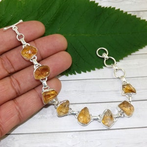 May include: A silver bracelet with a toggle clasp features seven faceted, orange-brown citrine stones. The bracelet is on a hand against a green leaf and wood background.