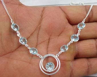 May include: A silver necklace with oval, pale blue gemstones. The necklace has a double-circle pendant with a single gemstone at the centre. The gemstones are linked by a fine silver chain. The necklace is displayed on a hand.