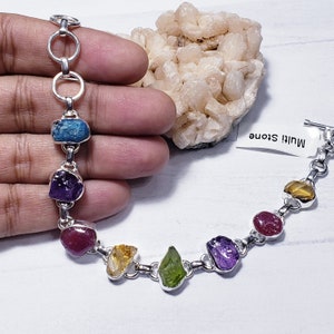 Op de afbeelding: Een zilveren armband met een kettingschakelontwerp en zeven verschillende gekleurde edelstenen. De edelstenen zijn blauw, paars, rood, geel en groen. De armband heeft een sluiting aan de rechterkant. Het label zegt "Multi Stone".