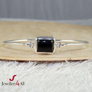 May include: A silver bangle bracelet with a square black gemstone. The bracelet is simple and elegant.