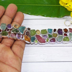 May include: A bracelet with a variety of colourful, irregularly shaped gemstones in shades of green, purple, red, and yellow. The stones are set in a silver-toned metal setting. The bracelet is approximately 18 cm long.