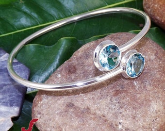 May include: A silver bangle bracelet with two blue topaz gemstones. The bracelet is open and has a simple design.