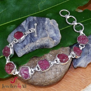 Könnte beinhalten: Ein silbernes Armband mit einem Schiebeverschluss zeigt sieben facettierte, rosafarbene Rubinsteine. Das Armband ist auf einem Hintergrund aus Stein und grünem Blatt fotografiert.