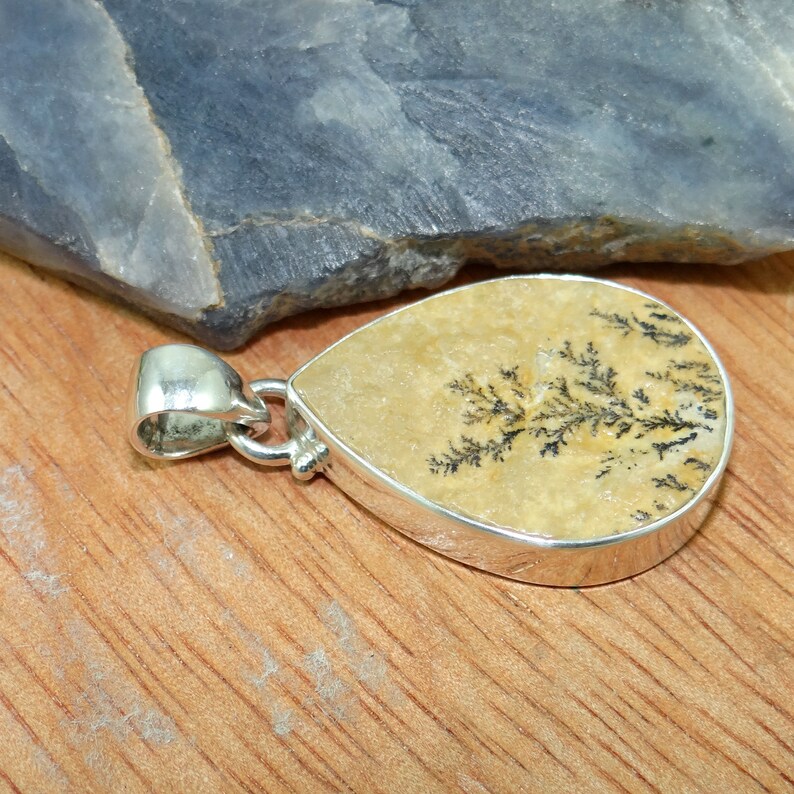 May include: A silver pendant featuring a teardrop-shaped piece of dendritic agate with a black and white tree-like pattern. The pendant is on a wooden surface.