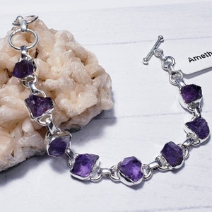 May include: A silver bracelet with raw, deep purple amethyst stones. The stones are irregularly shaped and set in a silver chain. The bracelet has a toggle clasp and a tag that reads "Ameth".