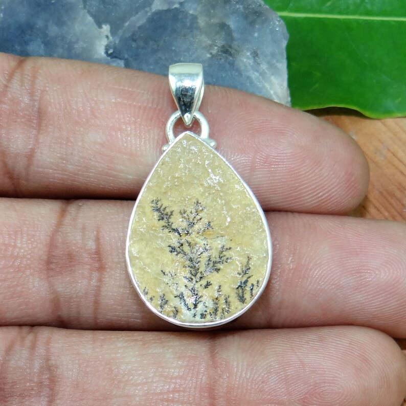 May include: A silver pendant featuring a teardrop-shaped stone with a dendritic agate pattern. The stone is a light beige color with black dendritic patterns.