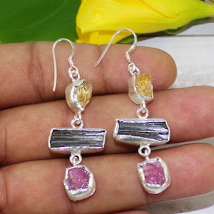 May include: A pair of silver earrings with a distinctive design. Each earring showcases a combination of raw gemstones: a yellow-orange stone, a dark grey rectangular stone, and a pink stone. The earrings are suspended from silver hooks.