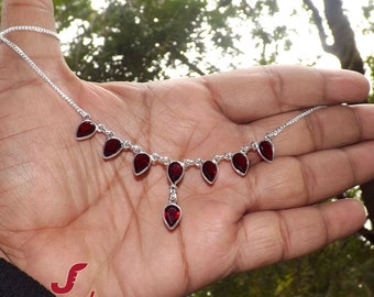 May include: A silver necklace featuring a series of teardrop-shaped, deep red gemstones. The gemstones are evenly spaced along the chain, with one pendant hanging lower. The necklace is displayed against a natural green background.