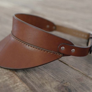 May include: A brown leather visor with a curved brim and adjustable strap. The visor is stitched with a lighter brown thread and has two silver rivets on the strap. The visor is designed to shield the eyes from the sun.