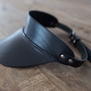 May include: A black leather visor featuring a curved brim and a thin adjustable strap that wraps around the head. The visor is constructed with visible stitching along the edge of the brim and is secured at the sides with small silver metal rivets.