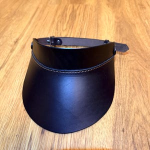 May include: A black leather sun visor featuring a wide, curved brim and a thin adjustable strap with a silver-toned metal buckle. The visor is constructed with visible brown stitching along the upper edge where the brim meets the headband.