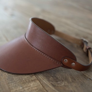 May include: A brown leather visor with a stitched seam and two silver rivets. The visor has a wide brim and a strap that goes around the back of the head.