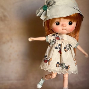 Puede incluir: Una muñeca con cabello rojo que lleva un vestido de estampado de Mickey Mouse verde claro y blanco, un sombrero verde claro con una flor y zapatillas verdes claras.