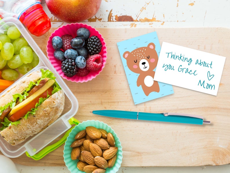 May include: A lunchbox with a sandwich, grapes, and almonds. A pink container with blueberries and raspberries. A blue and brown card with a cartoon bear and the text "Thinking about you Grace Mom".