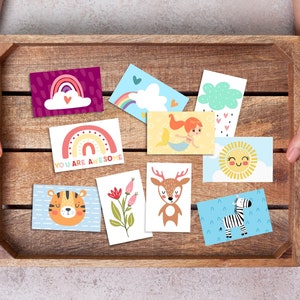 May include: A wooden crate filled with colorful cards featuring various illustrations, including a rainbow, a mermaid, a deer, a tiger, a zebra, a sun, and a cloud. The cards have different messages, including "You are awesome".