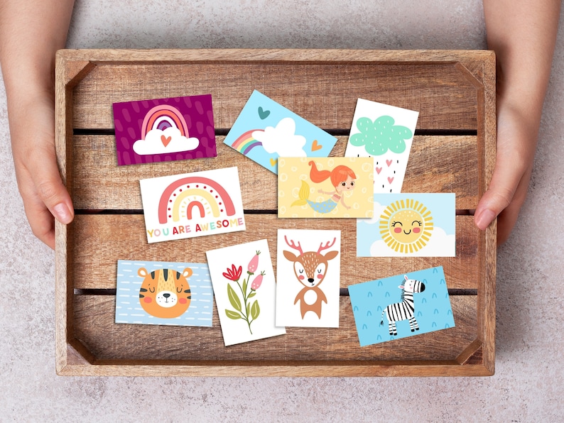 May include: A wooden crate filled with colorful cards featuring various illustrations, including a rainbow, a mermaid, a deer, a tiger, a zebra, a sun, and a cloud. The cards have different messages, including "You are awesome".
