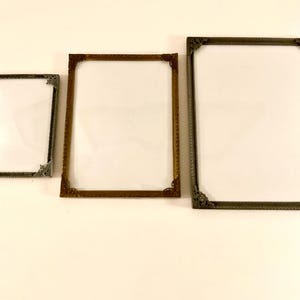 May include: Three rectangular picture frames with ornate corner details. The frames are in varying sizes and colors, including bronze and silver. The frames have a white interior, ready for photos or artwork.
