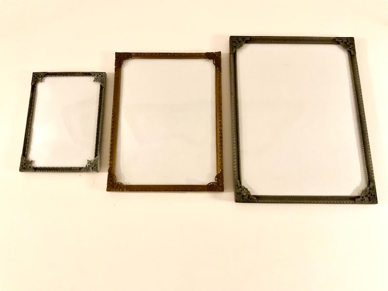 May include: Three rectangular picture frames with ornate corner details. The frames are in varying sizes and colors, including bronze and silver. The frames have a white interior, ready for photos or artwork.