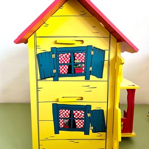 Op de afbeelding: Een geel houten poppenhuis met een rood dak en blauwe luiken. Het poppenhuis heeft twee ramen met rood-wit geblokte gordijnen en kleine bloempotten. Het ontwerp doet denken aan een sprookjesachtig huisje.