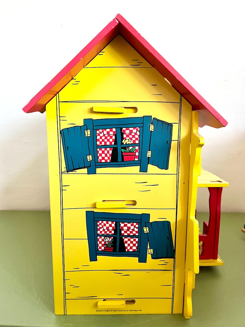 Op de afbeelding: Een geel houten poppenhuis met een rood dak en blauwe luiken. Het poppenhuis heeft twee ramen met rood-wit geblokte gordijnen en kleine bloempotten. Het ontwerp doet denken aan een sprookjesachtig huisje.
