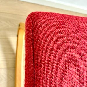 May include: Close-up of a red upholstered chair with a curved, light-coloured wooden frame. The chair's fabric has a textured appearance. The corner of the chair is visible, showing the rounded edge and the texture of the fabric.