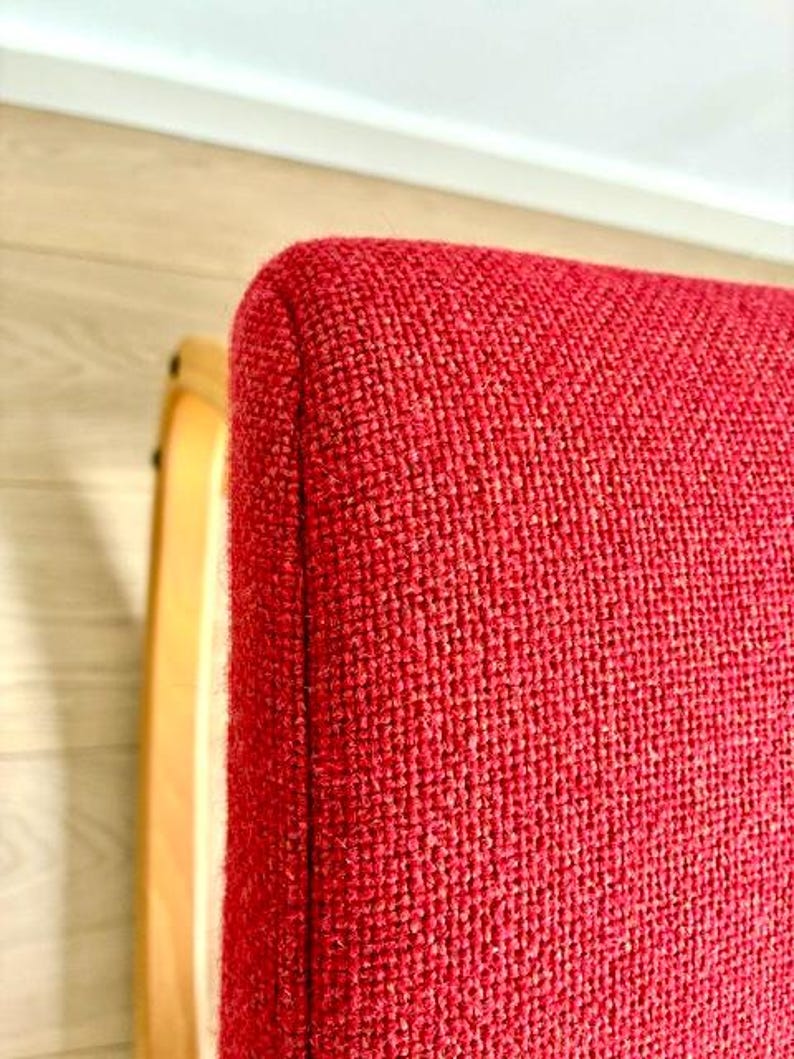 May include: Close-up of a red upholstered chair with a curved, light-coloured wooden frame. The chair's fabric has a textured appearance. The corner of the chair is visible, showing the rounded edge and the texture of the fabric.