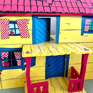 Op de afbeelding: Een kleurrijk houten poppenhuis met een gele buitenkant, een rood dak en blauwe deuren. Het poppenhuis heeft ramen met geblokte gordijnen en een kleine veranda met rode steunen. Het dak is gemaakt van rode tegels.