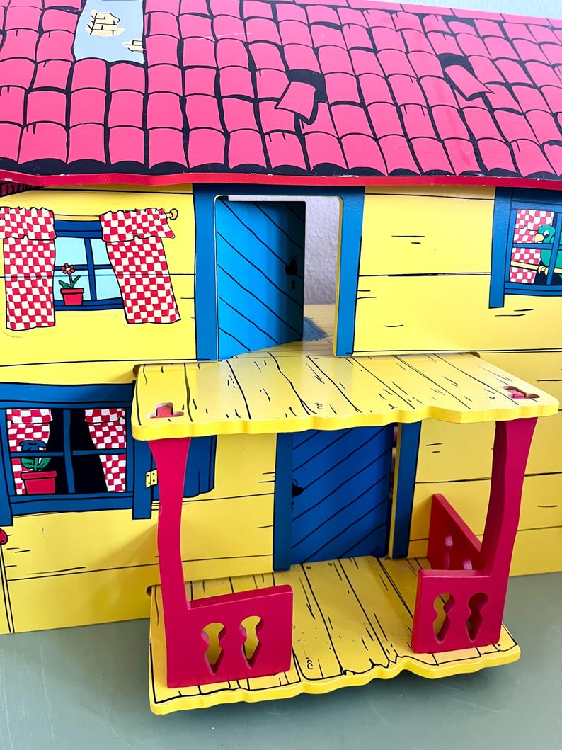 Op de afbeelding: Een kleurrijk houten poppenhuis met een gele buitenkant, een rood dak en blauwe deuren. Het poppenhuis heeft ramen met geblokte gordijnen en een kleine veranda met rode steunen. Het dak is gemaakt van rode tegels.