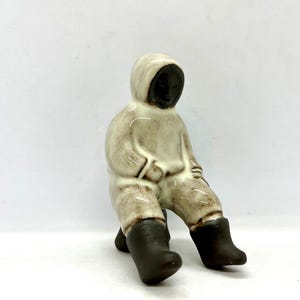 May include: A ceramic figurine depicting a seated figure in a hooded parka and boots. The figure's face is dark, contrasting with the off-white and brown tones of the clothing. The figurine is a decorative item.