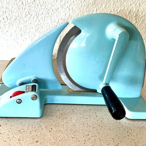 May include: A vintage, light blue meat slicer with a large, serrated blade. The slicer has a curved design, a black handle, and a red button. It sits on a light-colored surface.