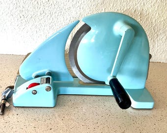 Pode incluir: Uma fatiadora de carne vintage azul claro com uma grande lâmina serrilhada. A fatiadora tem um design curvo, uma alça preta e um botão vermelho. Está sobre uma superfície de cor clara.