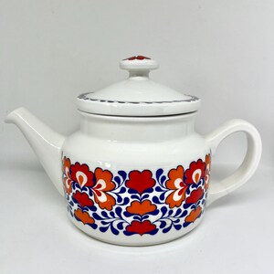 Puede incluir: Una tetera de cerámica blanca con un patrón floral repetitivo en tonos naranja, rojo y azul oscuro. La tetera incluye una tapa a juego con un pequeño pomo, un asa curva y un pico vertedor, todo decorado con un borde de color azul oscuro.