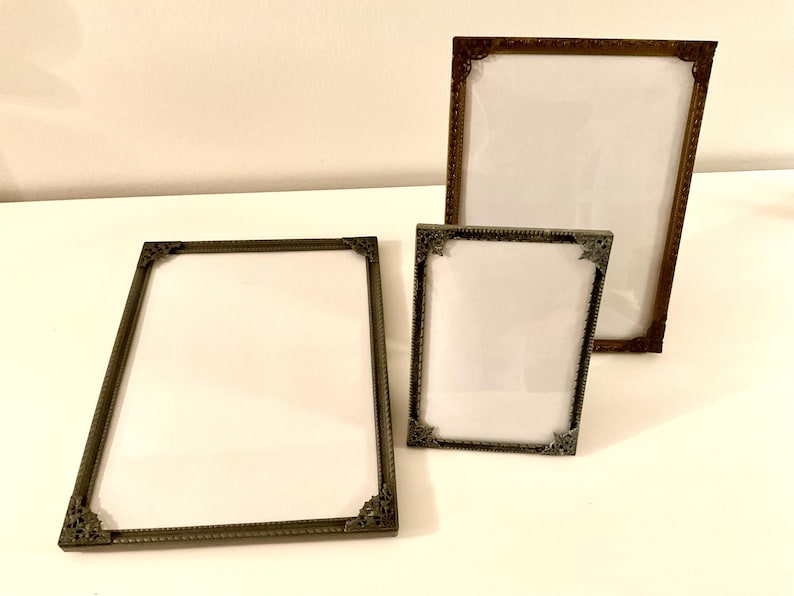 May include: Three antique picture frames with ornate corner details. The frames are in varying sizes and have a bronze-like finish. Each frame has a white backing, ready for a photo or artwork.