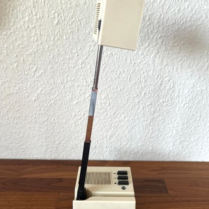 Könnte beinhalten: Eine weiße Schreibtischlampe mit einem schwarzen verstellbaren Arm und einem weißen Sockel. Die Lampe hat einen schwarzen Schalter und ein schwarzes Stromkabel.