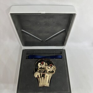May include: A gold-colored metal ornament in the shape of a flower basket, with two flowers and red gem accents. The ornament is displayed in an open white box with a dark gray interior and a blue ribbon.