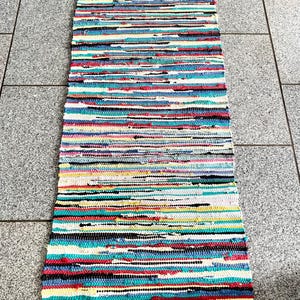 Puede incluir: Una alfombra rectangular multicolor de trapo con rayas horizontales. La alfombra presenta una variedad de colores, incluyendo rojo, azul, blanco, amarillo y negro. La alfombra tiene un borde con flecos en ambos extremos. Esta alfombra es adecuada para un pasillo o entrada.
