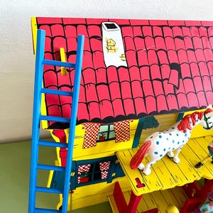 Op de afbeelding: Een kleurrijk houten speelgoedhuis met een rood dak, gele muren en een blauwe ladder. Een wit paard met blauwe stippen en rode manen staat op het dak. Het huis heeft een schoorsteen en kleine figuren.