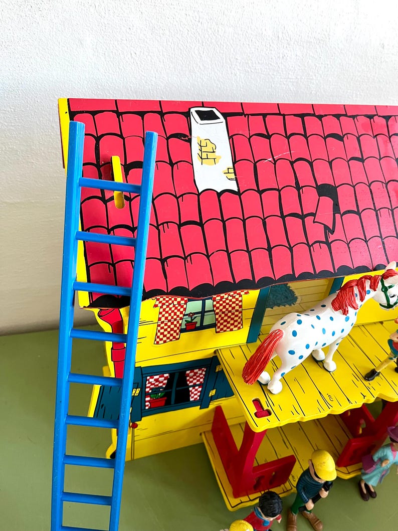 Op de afbeelding: Een kleurrijk houten speelgoedhuis met een rood dak, gele muren en een blauwe ladder. Een wit paard met blauwe stippen en rode manen staat op het dak. Het huis heeft een schoorsteen en kleine figuren.