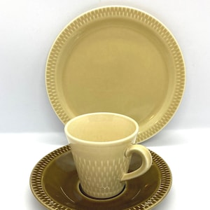 Puede incluir: Un juego de cerámica compuesto por una taza de color beige claro, un platillo marrón oscuro con borde texturizado y un plato a juego de color beige claro. La taza presenta un sutil patrón vertical, mientras que el platillo y el plato comparten un diseño geométrico en el borde.