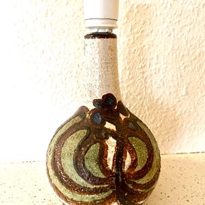 Könnte beinhalten: Eine Tischlampe aus Keramik mit weißer Lampenfassung. Der Lampenfuß hat eine bauchige Form mit einer strukturierten, cremefarbenen Oberfläche. Der Sockel ist mit braunen und grünen Wirbelmustern verziert. Ein weißes Stromkabel geht von der Basis aus.