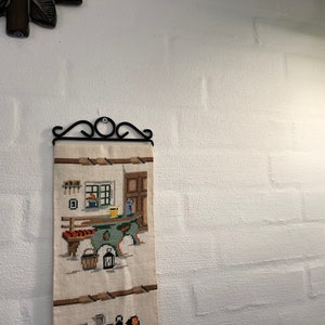 May include: A white wall with a black metal wall hanging featuring a beige tapestry with a cross-stitch design of a kitchen scene with a table, chairs, and a window.