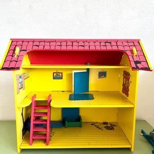 Op de afbeelding: Een felgekleurd houten poppenhuis met een rood dak, gele muren en een roze ladder. Het poppenhuis heeft twee verdiepingen met een blauwe deur en kleine ingelijste foto's aan de muren. Het dak is zwart afgewerkt.