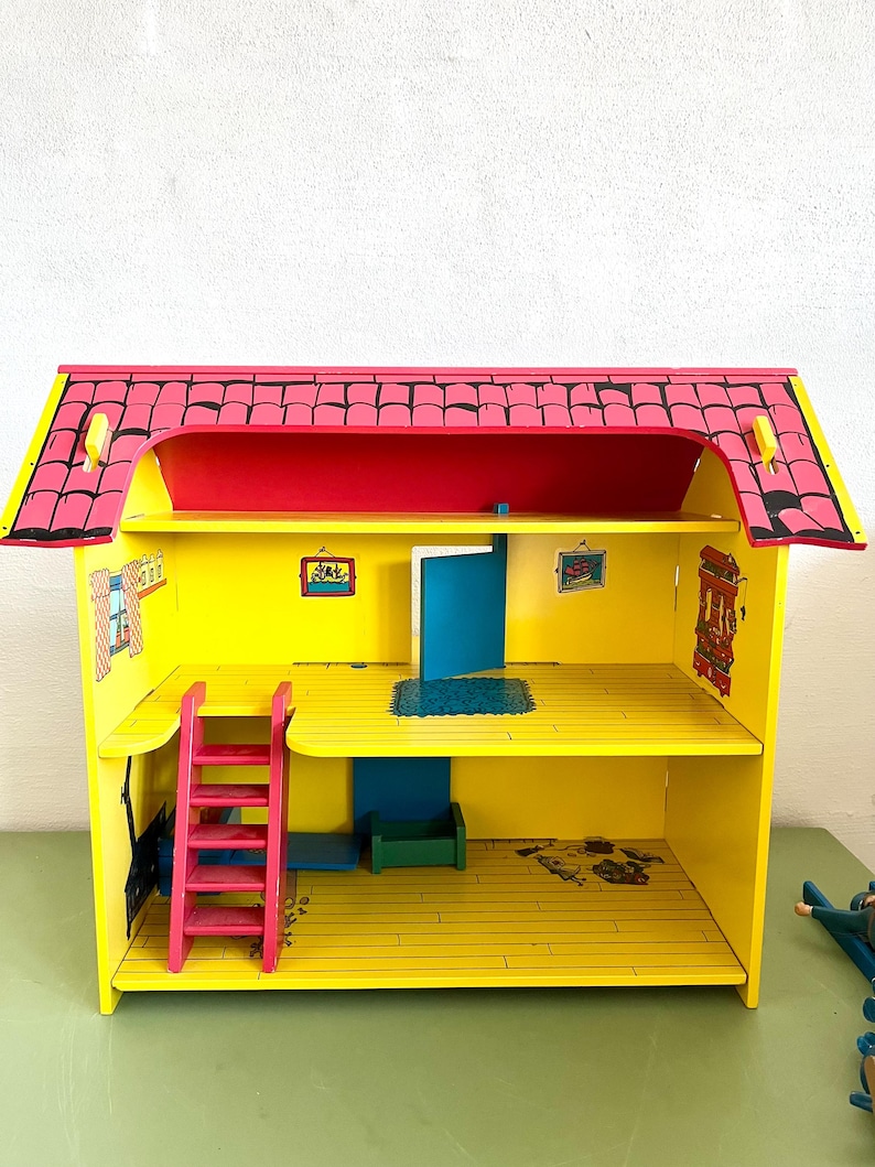 Op de afbeelding: Een felgekleurd houten poppenhuis met een rood dak, gele muren en een roze ladder. Het poppenhuis heeft twee verdiepingen met een blauwe deur en kleine ingelijste foto's aan de muren. Het dak is zwart afgewerkt.