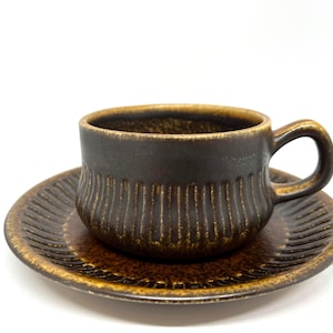May include: A brown ceramic teacup and saucer set. The cup has a handle and vertical ribbed detailing. The saucer also has ribbed detailing. The set is a dark brown color with lighter brown accents.