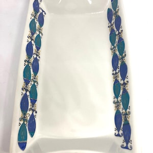 Può includere: Vassoio da portata rettangolare bianco con due manici. Il vassoio è decorato con un motivo ripetuto di forme a goccia blu e verde acqua con accenti dorati sui lati. Il vassoio è in ceramica.