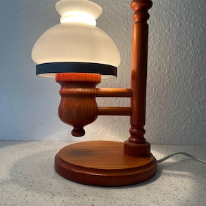 Może przedstawiać: Drewniana lampa stołowa z białym szklanym kloszem i czarną taśmą wokół klosza. Lampa ma drewnianą podstawę i drewniany wysięgnik, który rozciąga się, aby utrzymać klosz.