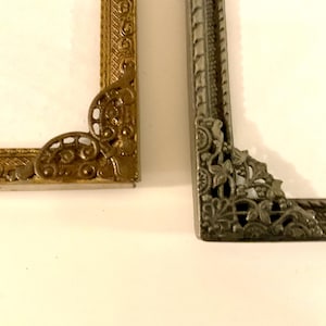 May include: Close-up of two ornate picture frame corners. One is gold-toned with scrollwork, the other is silver-toned with floral and leaf designs. Both frames have a white interior.