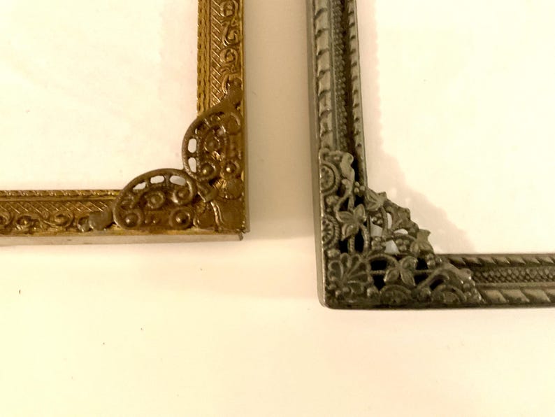 May include: Close-up of two ornate picture frame corners. One is gold-toned with scrollwork, the other is silver-toned with floral and leaf designs. Both frames have a white interior.